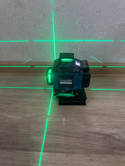 A green laser level projects intersecting horizontal and vertical lines onto two walls meeting at a corner. One wall has a textured, light-colored finish, possibly plaster or stucco, while the other wall features a wood-grain laminate. The laser level itself, a multi-faceted device with a black and teal casing, is mounted to the wood-grain wall. Visible text on the device reads "PATRON". The scene appears to be an indoor setting, likely a room or hallway, with no indication of the time of day or weather conditions. The activity is the projection of laser lines, suggesting a task related to construction, renovation, or measurement is in progress.
