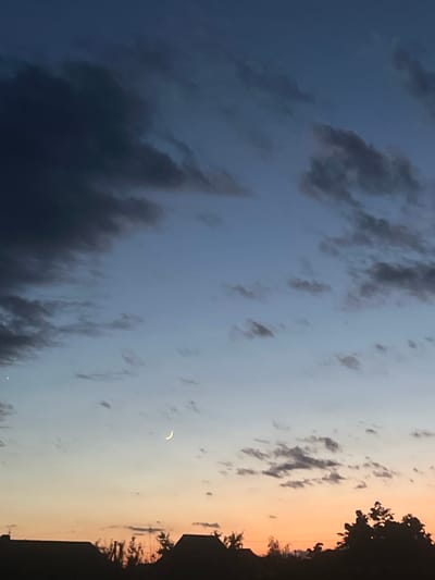 The image captures a dusky sky over the city of Slobozia, Moldova. The sky transitions from a deep blue at the top to a lighter blue and then to an orangey hue near the horizon. Scattered clouds, varying in shade from dark gray to lighter wisps, populate the sky. A sliver of a crescent moon is visible in the upper-middle portion of the frame. Silhouetted against the horizon are the rooftops of houses and the tops of trees, suggesting a residential area. There is no visible human presence or activity.