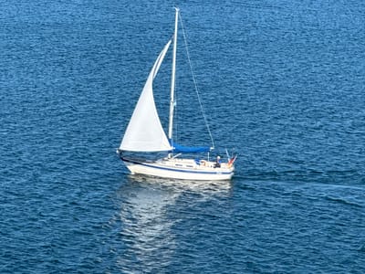 An aerial view captures a white sailboat with blue trim gliding across a deep blue, rippled body of water. The boat's main sail is fully deployed and catches the light, while a smaller blue sail is furled. A single person, wearing a light-colored hat, is visible on the deck of the sailboat, positioned near the stern. Two flags, one red and one with a yellow and blue design, are attached to the stern. The sailboat leaves a visible wake behind it, and its reflection is cast on the water's surface. The environment is entirely water, with no land features visible in the frame. This scene is consistent with the waters surrounding Karlskrona, Sweden, a coastal city known for its archipelago and naval history.