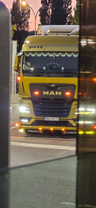 This is a medium shot, eye-level, outdoor photograph of a yellow MAN TGX truck. The truck is stationary and positioned at an angle, facing slightly to the left of the camera. The truck's headlights and orange indicator lights are illuminated. The license plate reads "BN-52KYO". The truck has "ROMANIA" decals with Romanian flags on either side of the lower front bumper. A teddy bear is visible sitting on the dashboard inside the cabin. The background shows trees and a street lamp with a pinkish-orange sky, suggesting it is dusk. The scene appears to be on a road or parking area. The image is partially obscured by a dark, reflective surface in the foreground, possibly a window or a car. The location is identified as the city of Vic, Spain.