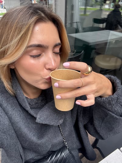 A young adult female is depicted in a close-up, chest-up shot, actively drinking from a plain, light brown paper cup. Her eyes are closed, and her lips are pursed towards the cup's rim. She possesses light brown hair with blonde highlights and a visible small gold stud earring in her left earlobe. Her right hand, featuring light pink manicured fingernails, holds the cup; a large, smooth gold dome ring is present on her right index finger, and a thin gold band ring on her right ring finger. She is dressed in a dark gray, textured sweater or coat, with a darker gray knit top visible beneath the wide collar. A black quilted leather handbag with a silver chain strap is slung over her left shoulder, resting on her lap. The background is a blurred reflection from a large glass window, indicating an outdoor urban environment beyond, with visible elements including green foliage, a light-colored building, and a dark, indistinct human silhouette walking leftward. Interior reflections show parts of minimalist white-topped tables and dark chairs, suggesting an indoor cafe or restaurant setting in Vaulx-en-Velin, France.