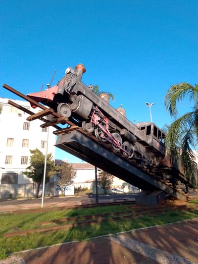 Located in Campo Grande, Brazil, the scene depicts a large, dark-colored steam locomotive sculpture angled dramatically upwards on a steel display structure. The historic-style locomotive, predominantly black with red-painted pilot and connecting rods, rests on a section of elevated railway tracks made of steel and wooden sleepers, ascending sharply towards the upper right. The entire structure is supported by a substantial dark-colored base. In the left background, a multi-story white building with rectangular windows and graffiti on its lower facade is partially obscured by trees. To the right, large green palm fronds partially frame the locomotive. The sky above is uniformly clear and blue. The foreground consists of a grassy area featuring exposed, derelict railway tracks, alongside a paved pathway exhibiting long, geometric shadows. A paved street runs parallel beyond the tracks, and a modern street light is visible on a pole in the upper right background. No people are present in the scene.