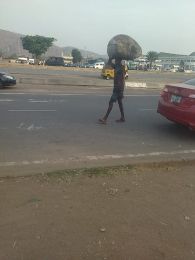 The scene takes place on a four-lane highway in Maraba, Nigeria. A man in dark clothing is walking across the highway, carrying a large, heavy object on his head. The object is covered in a gray tarp.  The man is walking away from the camera, towards the right side of the frame.  To the right of the man, a red car is stopped in the left lane of the highway.  To the man's left, there are multiple vehicles traveling in the opposite direction, including a yellow tricycle with a flatbed.  The highway is paved and has white lines painted on it. The ground on the side of the road is dirt.  There is a large, green tree in the background to the left of the road.  The sky is overcast and there is a low-lying hill in the distance.  A small, brown, grass area can be seen off to the left of the frame, and there is a large building in the distance on the right.  The man is the subject of the image, while the other objects are part of the background.