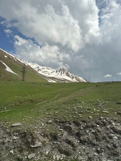 The image depicts a steep, grassy mountainside dotted with rocks and sparse vegetation. In the distance, a snow-capped mountain range rises dramatically against a cloudy sky. A lone, towering metal pylon stands prominently on the green slope, suggesting human infrastructure in this rugged landscape. The clouds are a mix of white and gray, hinting at overcast or partly cloudy weather. The lighting suggests it is daytime, possibly late morning or early afternoon, with the sun casting subtle shadows. The scene is characterized by its natural grandeur and the juxtaposition of elements like the green grass, white snow, rocky terrain, and the artificial structure of the pylon. No people or specific events are visible. The orientation of the image is rotated 90 degrees counter-clockwise, with the sky appearing on the left and the ground on the right.
