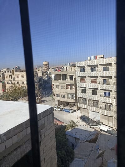 This image depicts an elevated view of an urban area in Mu'adammiyat ash-Sham, Syria, under a clear, bright blue sky.
In the immediate foreground on the left, a light-colored brick and concrete rooftop or balcony edge is partially visible, framed by a dark, vertical bar, likely part of a window or security grill from the vantage point of the photograph. Below this edge, sparse green foliage is present.
The mid-ground reveals a paved street running through a densely built environment. Along the street, several vehicles are parked: a blue sedan, a dark-colored SUV (possibly a Mercedes G-Wagen) with a utility trailer or attachment behind it, a white pickup truck, and another white van or small truck. No individuals are visible on the street or in the vehicles.
The street is flanked by multi-story residential and commercial buildings. On the right, a prominent building with a patterned facade of exposed concrete blocks features numerous windows, some with dark frames and others with horizontal slat blinds in reddish-brown. Balconies with dark metal railings are visible on its lower floors. The building directly opposite on the left side of the street is also multi-story, appearing older with some exposed concrete and what may be unfinished or damaged sections. Its rooftop supports several water tanks (red and white) and at least one satellite dish.
Further down the street, more buildings line the thoroughfare, receding into the background. A tall, light-colored cylindrical water tower with a conical top stands out prominently in the distant background, surrounded by additional buildings. The horizon line reveals low-lying, reddish-brown hills, suggesting a semi-arid landscape beyond the city. Several power lines and poles are visible throughout the urban landscape.