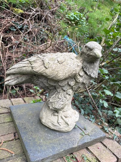 In Snailbeach, United Kingdom, a weathered light-grey stone statue of a bird, approximately 40 cm tall, stands prominently in an outdoor environment. The statue depicts a bird resembling an eagle or hawk, carved with textured feathering on its body and wings, with its head turned slightly right, facing forward. It rests on a rectangular dark grey stone slab, approximately 50 cm long and 30 cm wide, which exhibits minor moss growth and surface weathering. This slab is situated atop a ground surface composed of weathered, reddish-brown brick paving. The background is densely vegetated, featuring numerous intertwining bare brown branches, vibrant green ivy, other green foliage, and patches of moss. A segment of a blue rope or cord is partially visible amidst the background vegetation. No individuals are present, and no actions or interactions are depicted. The lighting indicates an overcast day or a shaded area.