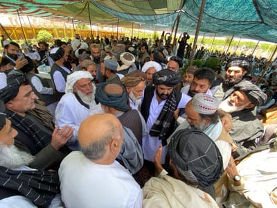 A gathering of men in a tent in Zaker-E Sharif, Afghanistan. The scene is dominated by a group of men in traditional Afghan clothing who are gathered around a man in a white turban and white robe. The man in the white turban appears to be talking to the group, and the other men are listening attentively. There is a large gathering of people surrounding the tent, and the men are all wearing turbans and traditional clothing. The men in the foreground are all dressed in white robes and turbans, and they appear to be engaged in a discussion. The environment is one of a religious gathering, with a large tent providing shade for the attendees. There are trees and a large gathering of people in the background. The scene is chaotic and crowded, with men of different ages and backgrounds gathering around the central figure.  It is likely that the men are observing a religious ceremony or celebration, and the man in the white turban may be a religious leader. The mood of the gathering is one of respect and reverence.