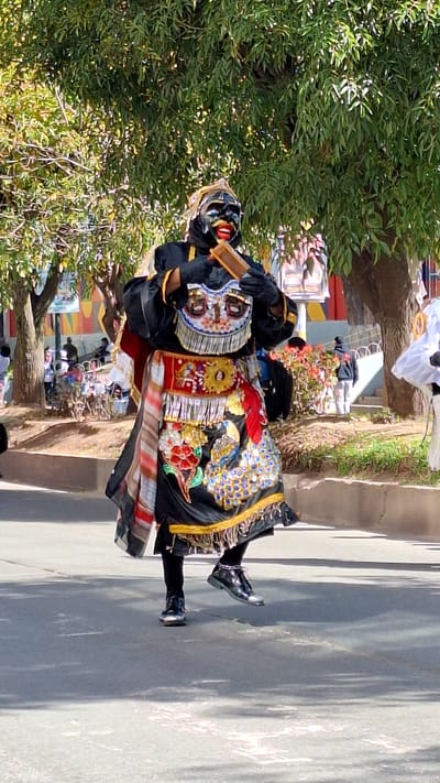 A performer in traditional attire with a black mask, red lips, and gold trim is dancing in the street in Wanchaq, Peru. The costume is black with intricate embroidery including a peacock design. The performer is wearing black gloves and shiny black shoes. The background features trees and buildings, suggesting an outdoor festival or parade.