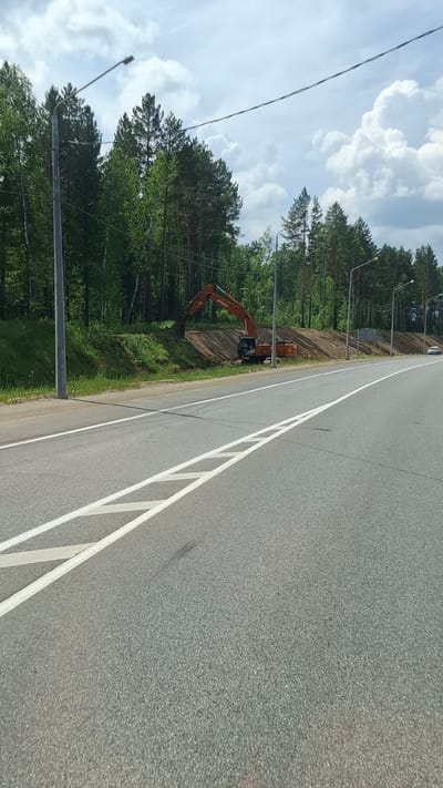 The image shows a highway construction site in the city of Moty, Russia. A large orange excavator is digging into a dirt embankment next to a newly paved road. The road has white lane markings. On the left side of the road, there is a tall grey pole with a street light attached, and beyond that, a dense forest of green trees. On the right side of the road, there are more street lights and a white car is visible in the distance. The sky is partly cloudy with white clouds against a blue sky.
