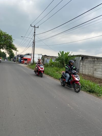 Two men are riding red motorcycles on a paved road in Kecamatan Margaasih, Indonesia. The road is lined with grass and trees. The sky is overcast with gray clouds. The men are both wearing helmets. One man is wearing a red and white jacket. The other man is wearing a light brown shirt. The motorcycles are both facing the same direction and are travelling on the right side of the road. The road appears to be empty of other traffic. There are multiple power lines overhead. The location is approximately -6.963 latitude and 107.538 longitude.