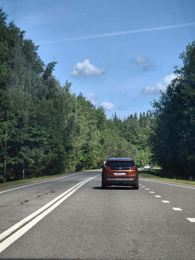 In this image, a brown Peugeot SUV is driving on a two-lane asphalt road. The car is in the right lane, heading away from the viewer. The road is lined by lush green trees on both sides, forming a dense forest. The sky is blue with scattered white clouds and a faint contrail. The road has white dashed lines marking the lanes and a solid white line on the right edge. The license plate of the Peugeot is visible and reads "9091 KT-2". In the distance, on the right side of the road, there are two other vehicles, a white car and a dark colored car, also driving away from the viewer. The scene depicts a typical rural road in Belarus, likely within the Юзуфоўскі сельскі Савет region, given the context.