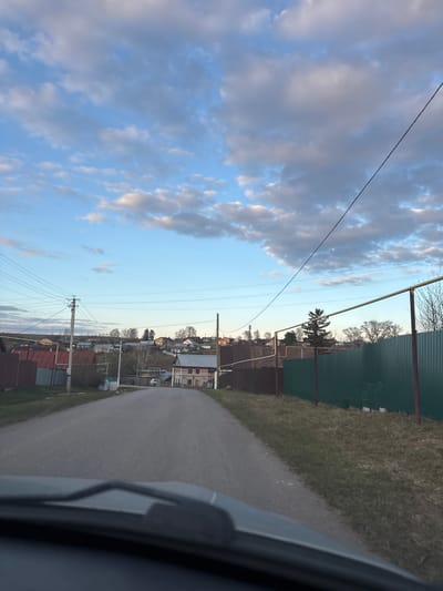 Captured from the interior of a vehicle, this image depicts a rural or semi-rural scene in Aktaziki, Russia, under a cloudy sky. The perspective is slightly angled, looking forward and to the right.

The upper portion of the image is dominated by a pale blue sky dotted with numerous scattered, large grey and white clouds, suggesting late afternoon or early evening lighting. Multiple thin power lines stretch across the sky, crisscrossing the frame, supported by several utility poles visible in the mid-ground.

In the foreground on the right, a paved asphalt road curves gently out of view. Adjacent to the road is a strip of dry, light green grass. Running along the right side of the frame, a tall, corrugated metal fence, painted dark green, parallels the road. Behind this fence, more grassy terrain is visible, along with a few bare trees and one distinct evergreen tree. A portion of a dark grey car interior or frame is visible on the extreme right, indicating the vantage point of the photograph.

The mid-ground and background reveal a scattering of houses and buildings, characteristic of a small village or settlement. These structures feature various rooflines and wall colors, with some appearing residential. Additional fences, some with reddish-brown panels, delineate properties. The overall atmosphere is quiet and observational, with no specific activity taking place. No text is visible in the image.