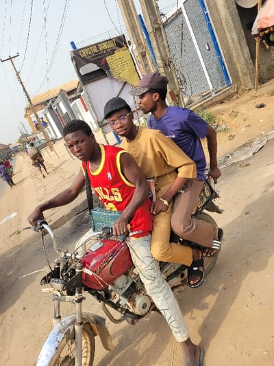 The scene depicts three young Black males riding a red motorcycle on an unpaved, dusty road in Puerto López, Colombia. The driver, positioned on the left, wears a red NBA Chicago Bulls jersey with "23" visible, light-colored patterned jeans, and dark flip-flops. He has a teal-colored messenger bag across his chest and looks directly at the camera while gripping the handlebars. The middle passenger, seated behind the driver, wears a light brown long-sleeved tunic, dark-rimmed glasses, a black flat cap, and light brown pants. He is looking towards his right. The rear passenger, seated behind the middle individual, wears a dark blue t-shirt, a camouflage baseball cap, brown pants, and dark Adidas slides with white stripes. He also looks to his right.
The motorcycle has a red "BOXER" branded fuel tank and shows signs of wear, including mud on the front fender. In the background, there are various buildings, including one with a "CRYSTAL HANGOUT" banner displaying text in yellow on a black background. Multiple utility poles with wires are visible. The sky is bright and appears hazy or partly cloudy. Faint figures of other individuals are discernible walking in the distance along the left side of the road.