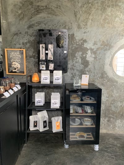 The image depicts an interior retail and display area within a cafe or coffee shop, situated in Kota Batu, Indonesia. The environment features dark, distressed concrete walls with prominent textures and cracks, a circular window on the upper right, and a dark concrete floor. No individuals are present in the scene.
On the left, a black counter displays multiple clear glass jars containing granular material, consistent with coffee beans or nuts, each labeled with small white tags.
Centrally, a black metal shelving unit is positioned, holding numerous light-colored bags, presumed to be packaged coffee products, distributed across its three shelves. Above this unit, a black pegboard is mounted on the wall, presenting a bronze-colored Buddha head sculpture centrally, flanked by various small merchandise items, such as keychains and accessories, suspended from hooks. To the immediate left of the pegboard, a framed artwork with a wooden border features a skull illustration and partially visible text including "life grows, death fades, coffee pending." A small, illuminated, orange-toned lamp is placed on the top shelf of the metal unit.
To the right of the shelving unit, a black, glass-fronted GEA refrigerated display case exhibits assorted food items on multiple shelves, including slices of cake (e.g., cheesecake, chocolate cake), pastries, and individually packaged snacks. The top surface of the display case holds a small clear glass bottle and a desk calendar displaying "14 Juli." The scene is static, showcasing the retail and display elements of the establishment.