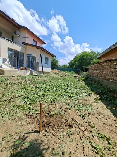 In Dolna Malina, Bulgaria, an exterior shot captures a residential construction site under a clear blue sky with scattered white clouds. In the foreground, a patch of freshly cut grass and weeds covers the uneven ground, with a wooden stake hammered into the dirt. A yellow string runs across the ground, possibly marking a boundary. To the left, a modern house with beige walls and a brown roof is visible, featuring large glass doors and windows, some of which are covered with protective plastic. An air conditioning unit is mounted on the exterior wall. Construction materials, including wooden pallets and white sheeting, are piled near the house's foundation. To the right, a brick wall with a concrete base forms a boundary, and a small wooden structure is visible behind it. Lush green trees and foliage border the property, suggesting a rural or semi-rural setting. The scene depicts an ongoing construction or landscaping project.
