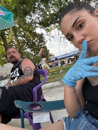 The image depicts an outdoor public gathering in Kostievo, Bulgaria. In the immediate foreground, a young adult female with dark hair, wearing a black tank top and denim shorts, is taking a selfie. Her right hand, covered by a light blue disposable glove, is positioned with the index finger to her lips in a "shhh" gesture. A silver charm bracelet, featuring what appears to be Olympic rings, is visible on her right wrist. She is seated in a low fabric camping chair with a built-in cup holder.
To her left, a middle-aged male with a beard and dark hair tied back is seated in a purple plastic chair. He wears a black sleeveless basketball-style jersey with white lettering and black shorts. His arms are heavily tattooed, with a distinctive detailed face on his right bicep and a serpentine or abstract design on his left forearm. He is looking towards the general direction of the camera. Behind him, a light-colored stone monument is visible, inscribed with Cyrillic text including "1300", "1981", and "БЪЛГА", consistent with a Bulgarian commemorative marker.
The background shows a public space with a mix of paved ground and grassy areas. An adult woman with blonde hair, dressed in a light-colored top and shorts, stands near a tree, looking to the right. Several children are present, including one in a white top and yellow shorts, playing on the grass. Further in the background, under multiple blue tarpaulin tents, a crowd of people is gathered, indicating a community event or market. The scene is illuminated by natural daylight, suggesting late afternoon or early evening. A green and white umbrella, partially visible in the upper left, displays "NESTLE" and "DREAM" branding.