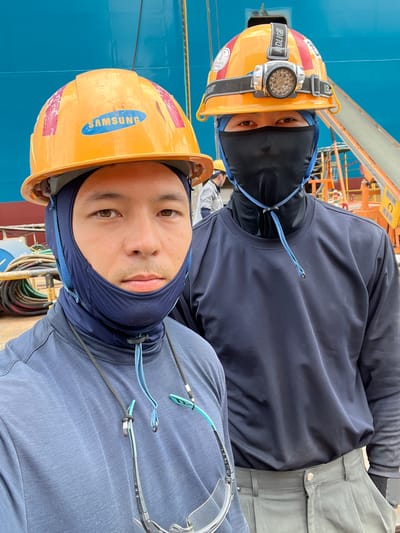 Two men, wearing hard hats and protective face coverings, pose for a selfie in front of a large blue structure, likely a ship or industrial building, with a backdrop of scaffolding and construction materials. The man in the foreground, on the left, wears an orange hard hat with a "SAMSUNG" logo and a navy blue neck gaiter pulled up over his face. He is wearing a grey long-sleeved shirt and clear safety glasses hanging from a lanyard. The man behind him, on the right, also wears an orange hard hat, this one equipped with a headlamp, and a black face covering. He is wearing a dark blue long-sleeved shirt and gray pants. The environment suggests an industrial work site, potentially a shipyard in Geoje-si, South Korea, given the prevalence of shipbuilding and related industries in the region. The lighting indicates an outdoor setting during daylight hours.
