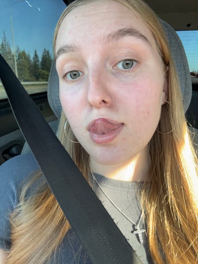 In Kukushtan, Russia, a young woman with blonde hair is seen from the chest up, seated in a vehicle. She is wearing a grey ribbed shirt and a silver cross necklace. Her tongue is sticking out of her mouth, and she has hoop earrings. A black seatbelt is diagonally across the image. The background shows a window with trees and the sky visible. The lighting suggests it is daytime, with sunlight casting shadows and highlights on her hair and face.