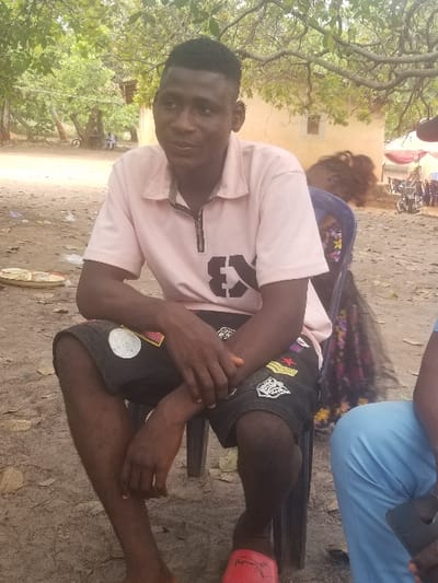 An outdoor scene in Ochadam, Nigeria, depicts a young adult male seated on a dark blue plastic chair. He wears a light pink short-sleeved polo shirt with a black zippered placket and a black "E3" graphic on the left chest. His lower attire consists of black shorts featuring multiple patches and designs. A red slide sandal with a white emblem is on his left foot; his right foot is bare. His hands are clasped in his lap, and he is looking slightly to his right. To the immediate right, a second individual is partially visible, wearing light blue trousers and holding a black mobile phone in their left hand. Further in the background, a third person with dark, curly hair is partially obscured. The ground is composed of dry earth with scattered leaves. In the mid-ground, a light-colored building with a rectangular window is visible. The far background includes green trees, a red canopy, several motorcycles, and two distant figures, one seated and one standing, near what appears to be a three-wheeled vehicle.