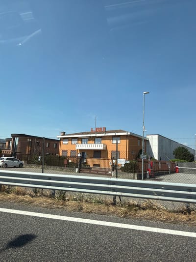 The image shows an orange building with a white balcony and a red sign with white lettering on the roof. To the left of the orange building is a darker building with a curved section. The buildings appear to be part of an industrial area in Cardano al Campo, Italy, as indicated by a sign visible on the orange building that reads "CARDANO AL CAMPO". There is a chain-link fence in front of the buildings, and a white car is parked to the left. A road with a white line is in the foreground, and a metal guardrail separates the road from the property. The sky is clear and blue. There are no people visible in the image.