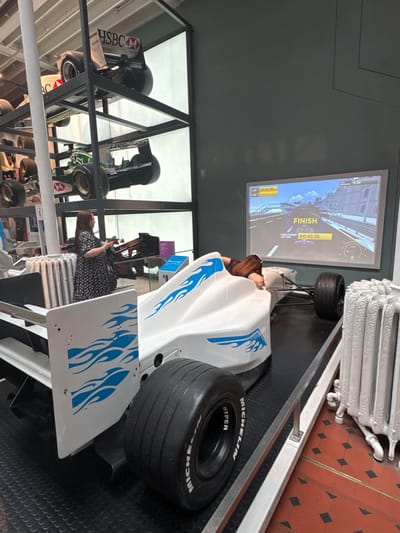 A woman, with long brown hair and wearing a black and white floral dress, stands to the left of a white and blue racing simulator in the City of Edinburgh, United Kingdom. The simulator, designed to resemble a Formula 1 car, is positioned on a black mat. Behind the simulator, a large screen displays a racing game, with the word "FINISH" prominently visible. To the left of the woman, several racing cars are displayed on tiered shelving units, some featuring sponsor logos such as HSBC and Bridgestone. A white, decorative radiator is located to the right of the simulator. The floor is a mosaic of red and black tiles.