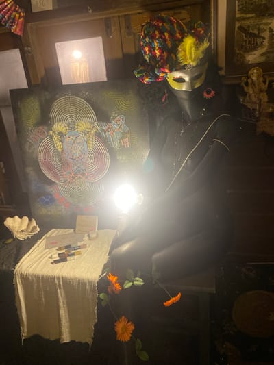 The image depicts an interior scene in what appears to be a shop or studio in Baan Don Sai, Thailand. The lighting is dim, and the view is from an elevated angle looking down. The most prominent elements include: a mannequin head wearing a colorful hat and mask; a large, dark, draped object resembling clothing or fabric; a canvas with intricate, colorful geometric designs; a stack of what appear to be paper or textile materials; and a few artificial orange flowers. There are also various smaller objects and art pieces visible in the periphery. The location is identified by a small note that reads "THE HANG @ TREE HOUSE" indicating that is likely the name of the location. The overall impression is one of a cluttered but creative space, possibly associated with art, fashion, or craftsmanship.