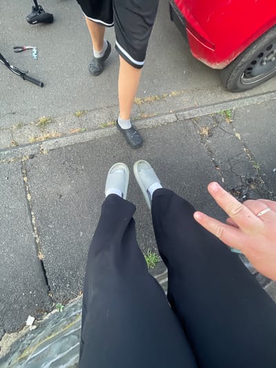 The image shows a ground-level view of two individuals standing on a paved surface, possibly a sidewalk or street, with a red car partially visible in the background. One individual is wearing black shorts and black Crocs with gray socks. The other individual is wearing black pants and light gray slides with white socks, and is making a peace sign with their right hand. There are also various objects scattered on the ground, including bicycle parts, a pair of pliers, and a small amount of grass and debris. The scene appears to be an outdoor urban environment. It is not possible to determine the specific location in Wiehl, Germany, from the provided image.