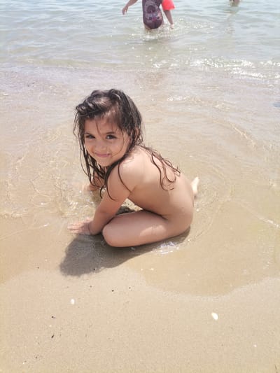 This media file shows a young girl sitting on the wet sand at the edge of the sea. The water is clear and calm, with small waves gently lapping at the shore. The girl is looking towards the camera with a happy and engaging expression. Her hair is dark and wet, falling around her face. She is wearing a dark swimsuit with a character on it and a bright orange arm float. In the background, another person is visible in the water, and the sun appears to be bright, creating a warm and inviting atmosphere. The scene is set at a beach, likely during the daytime, given the lighting conditions. The location is specified as Kranevo, Bulgaria. The overall mood is one of joy and relaxation, capturing a moment of a child enjoying a day at the beach.