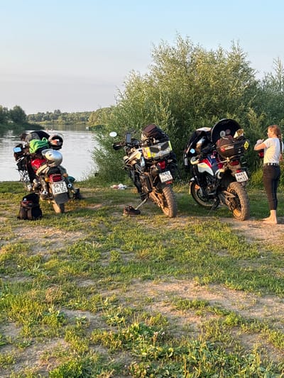 The image depicts a serene outdoor scene in Lan'shino, Russia. Three motorcycles, fully loaded with luggage, are parked on a grassy area next to a body of water, likely a river or lake. The motorcycles are oriented vertically in the frame, suggesting the original image was taken with a phone held in portrait mode and subsequently rotated.
In the background, a person is performing a plank exercise on the grass, facing away from the viewer. They are wearing a white t-shirt and dark pants, with their braid falling over their shoulder. The sun appears to be low in the sky, casting long shadows and illuminating the scene with a warm, golden light, indicating it's likely late afternoon or early evening. The weather seems pleasant, with clear skies.
The motorcycles are the primary objects of focus, each equipped with saddlebags, top cases, and other gear suitable for travel. One motorcycle has the license plate "6665 AM 55" and a sticker that reads "Motorider." Another visible license plate reads "5673 BH 50." The surrounding vegetation is lush and green, with a large tree providing shade. The water in the background is calm, reflecting the light and the trees along its edge. The overall mood of the image is one of peaceful exploration and outdoor activity.