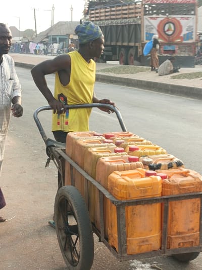 A man in a yellow tank top and blue head wrap pushes a two-wheeled cart loaded with numerous orange and yellow plastic containers. Another man stands beside him. The scene appears to be on a city street with a truck visible in the background. People are gathered on the side of the road, some sitting and working with piles of what seems to be dried leaves or grass. Buildings and structures are visible in the distance.