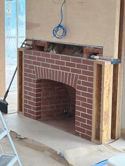 This media file captures a construction scene focused on a partially built brick fireplace in what appears to be a residential interior. The fireplace features a distinct arched opening and is constructed with red bricks laid in a stretcher bond pattern with white mortar. Above the brickwork, exposed wooden framing, likely OSB (Oriented Strand Board) and lumber, is visible, indicating the ongoing construction of the surrounding structure. A rolled-up bundle of blue electrical wire is loosely coiled on a plywood surface to the left of the fireplace. To the right, a large sheet of brown paper, possibly for protection, is draped over a section of the construction, with some text and graphics visible on its surface. A broom with a long handle is leaning against a window in the lower part of the frame, suggesting that cleaning or site preparation is underway. The lighting suggests it is daytime, likely indoors, with natural light coming from a window. The scene is devoid of people but implies human activity through the presence of construction materials and tools. The overall impression is one of a home renovation or new build project in progress.