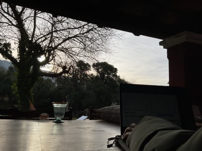 The image captures a view from a covered outdoor patio, looking out towards a tree-lined landscape at dusk. In the foreground, a wooden table is partially visible, with a glass and a laptop placed on it. A person's arm and hand are resting on the laptop, suggesting they are actively using it. The laptop screen displays text, but it is not clearly legible. Behind the table, a large, bare tree dominates the left side of the frame, its branches silhouetted against the pale sky. The sky itself is overcast with hints of warm light breaking through. The background consists of dense foliage and distant hills, indicative of a rural or semi-rural setting. The overall atmosphere is calm and somewhat muted, with the low light conditions casting long shadows. The location is identified as El Rodeo, Argentina.