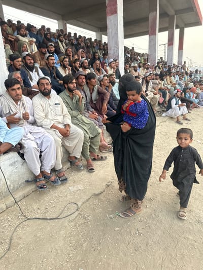 This image depicts a large gathering of people, predominantly men, seated and lying on the ground outdoors. The scene is set in a dusty, open area next to a concrete structure with white and red horizontal beams. Several individuals are wearing traditional South Asian attire, including shalwar kameez, some with white caps or turbans.
In the foreground, a woman in a black abaya is holding a young child wearing a blue shirt with red trim. To her right, a young boy in a black outfit is standing and looking towards the camera. The presence of children suggests a community gathering. The people appear to be looking in various directions, some upwards, as if observing something or listening to an announcement. Their expressions are neutral or slightly weary, with no clear indication of distress or celebration.
The ground is sandy and littered with small debris. A black cable snakes across the ground, its purpose unclear. The lighting suggests it is daytime, likely overcast or hazy, as there are no strong shadows. The context provided places the location in Chaman City Tehsil, Pakistan. The overall impression is of a communal event or a gathering for a specific purpose within this Pakistani locale.