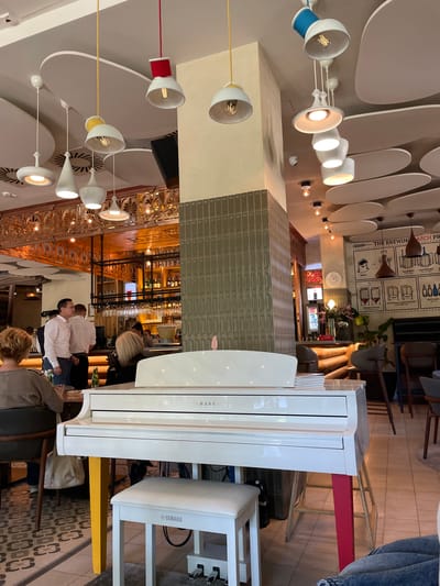 This image depicts the interior of a modern and stylish restaurant or bar, likely in Subotica, Serbia. The scene is brightly lit, showcasing a lively atmosphere with several patrons and staff members present.
In the foreground, a pristine white Yamaha grand piano with a matching bench dominates the space, with a person seated at the keys, suggesting live music or entertainment. Guests are seen socializing, some seated at tables with drinks, while others stand near a well-stocked bar area in the background, where staff in white shirts are visible.
The establishment features a contemporary design with notable details. The high ceiling is adorned with white, oval acoustic panels and an array of distinctive pendant light fixtures, some featuring vibrant blue, red, and yellow accents around their bases or cords. A prominent wall section is covered in sleek, grey rectangular tiles. A large, informative wall poster details "THE BREWING PROCESS," illustrating stages such as "MALT," "MASHING," "LAUTERING," "FERMENTATION," "MATURING/CONDITIONING," and "BOTTLING," indicating a craft beer or brewing focus. Other visible text includes "YAMAHA" on the piano and bench. The floor is tiled with an intricate, repeating geometric pattern, contributing to the sophisticated aesthetic. The overall impression is one of a chic, welcoming venue.