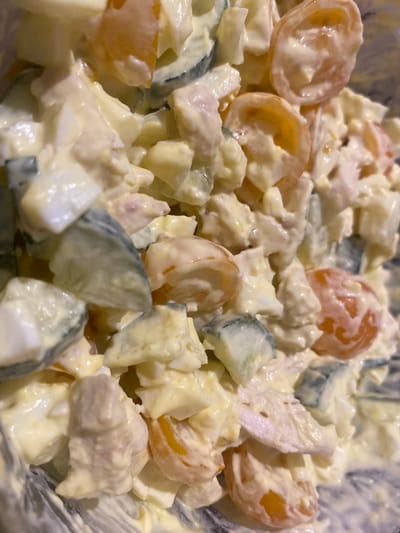 The image is a close-up, overhead shot of a bowl of salad. The salad appears to be made with chopped hard-boiled eggs, sliced tomatoes, and pieces of cucumber, all coated in a creamy white dressing, likely mayonnaise. The lighting is artificial and somewhat dim, casting soft shadows on the ingredients. The focus is sharp on the foreground ingredients, with some background blur. The salad is presented in a bowl that has a slightly textured, dark gray interior.
The salad consists of irregular, bite-sized pieces of the ingredients. The eggs are white and yellow, the tomatoes are a pale orange-red, and the cucumber pieces show a distinct green rind and lighter green flesh. The dressing is thick and opaque, coating everything evenly. There are no people visible in the image, and no specific setting is discernible beyond the bowl itself.
The image does not depict any particular event or activity beyond the presentation of the food. There are no indications of time of day, weather, or specific location cues, other than the provided context of Kolomna, Russia. The overall mood of the image is neutral, focusing solely on the food item. No text is visible in the image.