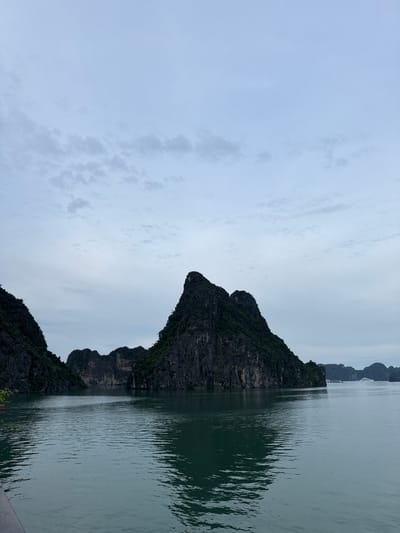 A wide shot captures the limestone karsts of Ha Long Bay, Vietnam, a UNESCO World Heritage site located within the administrative boundaries of the city of Hai Phong. The karsts, covered in lush green vegetation, rise dramatically from the calm, teal-colored waters of the bay. The sky above is overcast with a pale blue hue, with wispy clouds scattered across it, suggesting either early morning or late afternoon. In the distance, a few tour boats are visible, adding a sense of scale and hinting at the bay's popularity as a tourist destination. The water reflects the muted light and the silhouettes of the karsts, creating a serene and somewhat dramatic scene. The overall impression is one of natural beauty and geological grandeur.