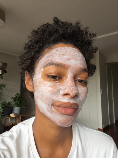 A close-up, head-on shot shows a young Black person with curly, dark hair, wearing a white t-shirt and a white face mask. The mask covers most of their face, leaving their eyes, nose, and lips visible. The person is looking directly at the camera with a slight smile. The background is blurred but appears to be an indoor setting with a light-colored wall and some plants on a shelf to the left. The image is well-lit, with natural light coming from the right.