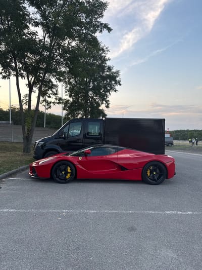 The scene depicts a red Ferrari LaFerrari hypercar parked on an asphalt surface with faded white parking lines, positioned in what appears to be a parking or service area of the Hungaroring circuit in Mogyoród, Hungary. The vehicle is oriented facing left, showcasing its full profile, featuring black multi-spoke wheels and prominent yellow brake calipers.

Behind and to the left of the Ferrari, a black Mercedes-Benz Sprinter van with a matching black box body is parked. Several trees with dense green foliage are situated behind the van, obscuring part of a concrete wall that runs horizontally across the mid-ground.

The background, particularly to the right, reveals an open, possibly unpaved area where approximately 3-5 indistinct human figures are standing in a small group, accompanied by another light-colored van. A distinctive red and white striped structure, indicative of motorsport track infrastructure, is also visible in this distant section. The sky overhead is a clear light blue with scattered wispy white clouds, suggesting a daytime setting with soft, ambient light. All vehicles are stationary, and no specific actions or interactions are discernible in the immediate foreground.