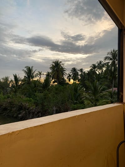 The image depicts a view from a building in Tanjongkapai, Indonesia. Through a window or opening, a tropical landscape is visible. Palm trees dominate the scene, silhouetted against a sky filled with clouds. The light suggests either sunrise or sunset. The building's structure, with its beige-colored wall, frames the external view.