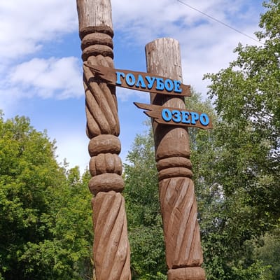 This media file depicts an outdoor scene featuring two intricately carved wooden posts, likely part of an entrance or monument. The posts are tall and weathered, with spiral and geometric carvings, suggesting a rustic or traditional aesthetic. Attached to the posts are two wooden signs with blue lettering. The top sign reads "ГОЛУБОЕ" and the bottom sign reads "ОЗЕРО". These words translate to "Blue" and "Lake" respectively.

The background consists of lush green trees, indicating a natural setting, possibly a forest or park. Above the trees, a bright blue sky with scattered white clouds is visible, suggesting fair weather and daytime. A thin wire stretches across the top of the frame, hinting at the presence of nearby infrastructure.

There are no people visible in the frame. The overall mood is serene and natural, with the carved wooden elements adding a unique artistic touch to the landscape. The location is identified as Sdt Shcherbakovka, Russia, which aligns with the Cyrillic text on the signs. The scene is well-lit, suggesting it was captured during daylight hours, likely midday or afternoon.