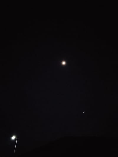 The image shows a dark night sky with two celestial objects and a street light. The largest celestial object, likely the moon, is in the upper center of the frame, appearing as a bright, somewhat blurred orb. To the right and lower than the moon, a smaller, distinct point of light, possibly a planet or star, is visible. In the lower left of the image, a street light with a white bulb is illuminating a thin, vertical pole. The silhouette of what appears to be a building or structure is visible at the very bottom of the frame, against the dark sky. The scene is set at night in Rumuokwuta, Nigeria, with the artificial light from the street lamp contrasting with the natural light of the celestial bodies.