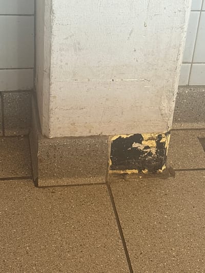 The image shows a close-up view of a corner where a white, concrete-like beam meets a textured, speckled grey wall. The beam is on the left, and a portion of it is painted a light color, possibly white or a pale gray. The wall on the right is made of rectangular panels with a grainy, speckled texture, appearing to be made of concrete or a similar material.
In the corner where the beam and wall meet, there is an area where the surface material is damaged and peeling. A dark surface, possibly black, is visible underneath layers of yellow and white material that are flaking off. This damage creates an uneven, textured patch in the corner. The lighting suggests an indoor environment, possibly a public space like a restroom or hallway, given the tiled elements visible in the background on the far left and the robust construction materials. There is no visible text in the image. The overall impression is one of wear and tear in a utilitarian architectural setting.