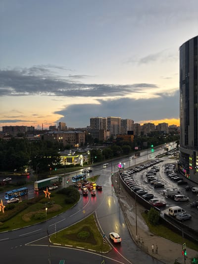 This is a high-angle, wide-shot photograph taken at dusk in Tarasovka, Russia, capturing an urban street scene after a rainfall. The sky is a gradient of pale blue to soft orange and yellow, with streaks of gray clouds. The wet asphalt reflects the light from street lamps and car headlights, creating a slick, shimmering surface on the road.
In the foreground, to the right, is a modern, multi-story glass-fronted building with its illuminated facade displaying various signs. Adjacent to this building is a large, curved parking lot filled with a dense arrangement of parked cars, primarily sedans and SUVs. The parking lot curves towards the center of the image.
To the left of the parking lot, a multi-lane road curves around a landscaped island. This island is adorned with large, illuminated star-shaped decorations. A blue city bus and a green semi-trailer truck with the "ITECО" logo are visible on the road, along with several other cars. The bus is positioned to the left, while the truck is further down the road. Traffic lights are visible, some showing red and green signals.
Further back, the cityscape unfolds with numerous apartment buildings and other structures of varying heights. A gas station with bright yellow canopies is visible on the left side of the road. Trees line the areas between buildings, creating a contrast between the urban development and pockets of greenery. The overall impression is a busy urban environment at the end of the day, with signs of recent precipitation adding a reflective quality to the scene. Two pedestrians are visible on the sidewalk in the lower right portion of the image, near the parking lot.