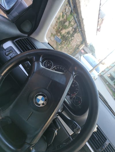 The image captures the interior of a car, specifically the driver's side dashboard and steering wheel. The steering wheel is black leather with a BMW logo in the center. Part of the car's instrument cluster is visible, showing a speedometer and a tachometer. A car key is hanging from the ignition. Outside the car, through the windshield, a street scene is visible. Buildings and foliage can be seen, suggesting an urban or semi-urban environment. The sunlight is bright, casting a glare on the windshield and parts of the interior. The location is specified as Madan, Bulgaria.