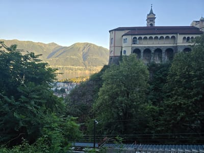 A wide-angle, high-angle exterior shot captures a scenic view from Orselina, Switzerland, featuring a building with distinctive architecture overlooking a lake and surrounding mountains. The building, appearing to be a religious or historical structure, is situated on a cliff, with its facade adorned with arched windows and a clock tower. Lush green trees in the foreground frame the scene, leading down to a body of water reflecting the sunlight and a town nestled along its shore. The mountains in the background are covered in dense vegetation, their slopes sculpted by the late afternoon or early morning sun, creating a dramatic play of light and shadow. The sky above is a clear, pale blue, indicating fair weather. The image is composed of natural elements, with no human figures or animals visible. The overall impression is one of serene natural beauty and architectural grandeur.