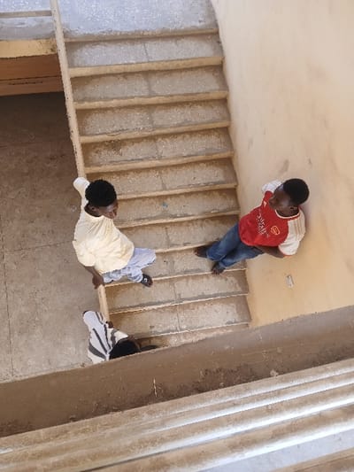 This image, taken from an elevated perspective, depicts a concrete staircase and an associated landing area within a building in Ugi Majowel, Nigeria. Three individuals are present in the scene. A dark-skinned male subject, wearing a light beige long-sleeved shirt and light grey trousers, stands barefoot on a concrete landing, looking upwards. To his right and positioned higher on the staircase, a second dark-skinned male subject, wearing a red and white short-sleeved shirt with "Emirates Fly Better" branding and blue jeans, leans against a plain beige wall; this individual is also barefoot. A third individual, partially visible from the head and shoulders, is positioned below the bottom landing, wearing a white shirt with black horizontal stripes. The concrete steps and landings show signs of wear and discoloration. A light-colored banister or handrail runs along the left side of the staircase.