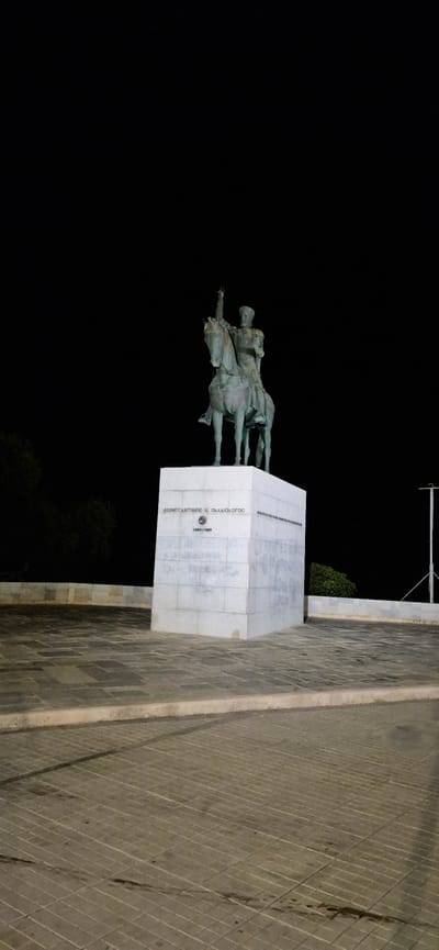 An equestrian statue of Constantine XI Palaiologos stands prominently on a marble pedestal at night in Paleo Faliro, Greece. The statue depicts the Byzantine emperor in military attire, with his right arm raised in a gesture of command. The dark night sky serves as a stark backdrop, with the illumination focused on the monument, creating dramatic shadows. The pedestal is inscribed with Greek text and what appears to be a circular emblem. The ground in front of the statue is paved with square tiles, and a stone wall with a textured surface runs along the perimeter. Trees are visible in the dark background on the left, and a modern, minimalist lamppost stands to the right. The overall scene conveys a sense of historical significance and solemnity.