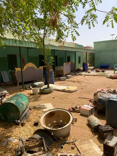 The image shows a scene of apparent destruction and disarray in an open courtyard in Khartoum, Sudan. The coordinates provided, latitude 15.552 and longitude 32.57, roughly correspond to a location in the city. The image shows a two-story building with a covered porch in the background, likely a residence or business, with a green painted exterior. The courtyard is littered with debris, including broken furniture, overturned barrels, tires, and a large metal rim.  Two mattresses are lying on the ground. The scene appears abandoned and neglected. There are no people present in the scene.  The presence of debris and abandoned objects suggests a recent event of disturbance or conflict.  The building appears to be damaged with a missing door. The green paint on the building shows signs of chipping. The courtyard is covered in a layer of dirt and dust, indicating a lack of recent maintenance.  The area behind the building is partially obscured by the trees, but a second structure can be seen. The trees in the background provide shade and some greenery to the scene. There are no signs of recent activity, except for the debris left behind. The environment appears dusty and dry, with the ground covered in a layer of sand and loose debris.