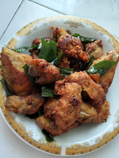 Golden-brown fried chicken pieces are arranged in a white bowl with a decorative gold rim. Interspersed among the chicken are vibrant green leaves and sliced green stalks, possibly herbs or garnish. The chicken appears crispy on the outside, hinting at a savory and aromatic dish. The presentation suggests a home-cooked meal.