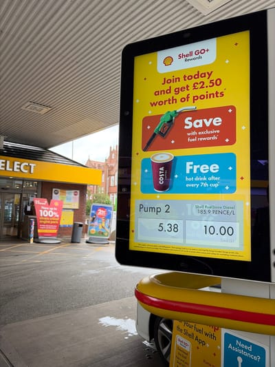 A digital screen attached to a fuel pump at a Shell petrol station in Leicester, United Kingdom, is the central focus of the image. The screen displays various advertisements and transaction details.
On the left side of the screen, there's a promotion for "Shell GO+ Rewards," encouraging users to "Join today and get £2.50 worth of points" and "Save with exclusive fuel rewards." Below this, a green fuel nozzle is depicted. In the center, a "Costa Coffee" logo is featured alongside an offer for a "Free hot drink after every 7th cup." The right side of the screen shows details for "Pump 2," indicating "Shell FuelSave Diesel" priced at "185.9 PENCE/L." The transaction shows "5.38 Litres" dispensed, totaling "10.00" pounds. Below these figures, small text outlines "T&Cs apply," directing users to www.shell.co.uk/shellgoplustc and detailing conditions for points.
To the right of the screen, part of a yellow and red fuel pump pipe is visible, along with a glimpse of a car wheel. Further right, an additional sign promotes "your fuel with the Shell App," urging users to "Download the Shell App and join Shell GO+," and a "Need Assistance?" message.
In the background, a Shell Select convenience store is visible, with part of its yellow signage reading "ELECT." Another sign nearby advertises Shell V-Power, stating, "Now up to 100% cleans critical engine parts." The ground is paved, and a distant brick building with traditional architecture can be seen under a bright, possibly overcast, daytime sky. The overall scene depicts a typical petrol station environment.